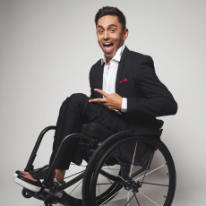 Danny J. Gomez in a suit sits in a wheelchair, smiling and holding up a peace sign. 