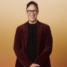 John Hoffman in a maroon velvet jacket and black shirt, smiling against an orange background.