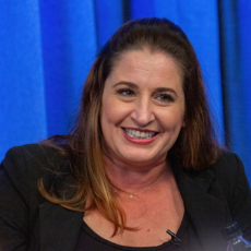 Kelli McNeil-Yellen smiling during a discussion at an event, with a blue background.