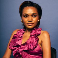 A headshot of Lolo Spencer in a pink riffle dress against a light navy background. 
