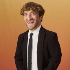 Nic Novicki in a black suit and tie against an orange background.