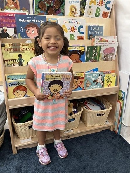 A little girl in a grey and white striped dress holding a book in front of a bookcase in a classroom.