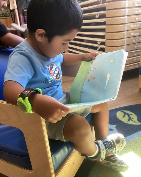 Jameson in a classroom sitting in a chair reading a children's book. 