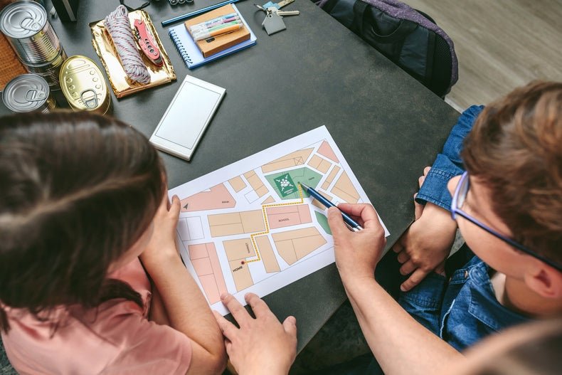 Kids sitting at a table in the living room surrounded by emergency kit supplies while an adult is showing them a map. 