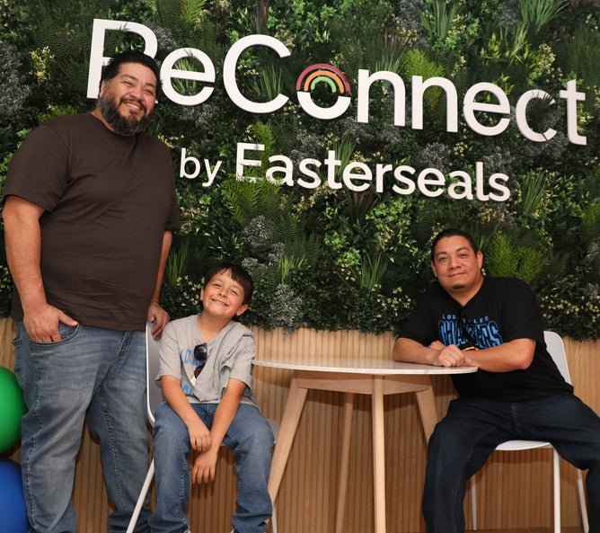 Three individuals in the new Sensory Space room sitting a table in front of a large moss wall.