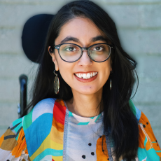 Shaina Ghuraya a smiling person with glasses, wearing colorful clothing, against a gray brick background.
