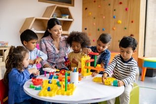 Adult and children engaged in building toys at a table in a colorful classroom setting.