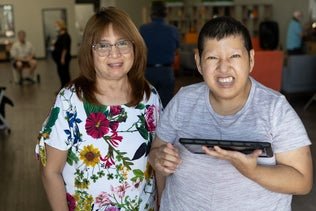 Two individuals smiling at the camera, one holding a tablet, in a community center setting.