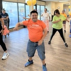 A cheerful group of people participating in a dance class at a community center. The person in the foreground wears an orange t-shirt with the text "WE CELEBRATE DIVERSITY AND DISABILITY" and denim shorts. Others in colorful attire are visible in the background.