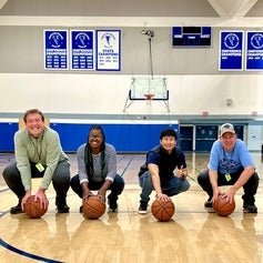 Four adults crouching in a gymnasium, each holding a basketball, with banners displaying "Championship" hanging on the wall behind them.