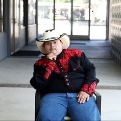 Person in a cowboy hat and red embroidered shirt sitting on a chair in a corridor.