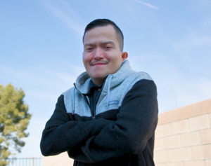Person wearing a gray hoodie with arms crossed, smiling against a blue sky background.