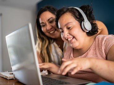 young girl with disability wearing headphones and using a laptop next to adult woman smiling