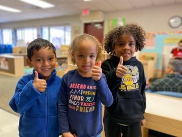 Three kids give thumb's up to the camera while standing in a Head Start classroom