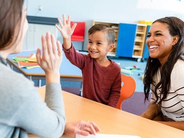 A young child high-fives a teacher across a table in a brightly colored classroom, while another adult smiles beside him, showing an encouraging and cheerful atmosphere.