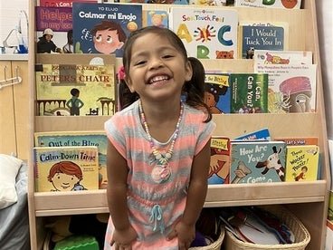 A little girl in a grey and white striped dress posing in front of a bookcase in a classroom.