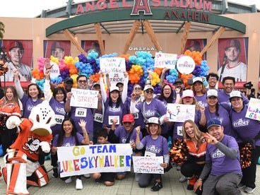 A group of individuals wearing purple Strides t-shirts and holding up posters in front of a balloon arch at the Angel Stadium. 