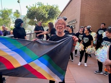 A person holding a Disability Pride flag smiles broadly in an outdoor setting with a cheerleading group in the background.