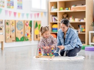 An adult and a child are sitting on the floor, engaging with colorful building blocks in a bright, art-decorated classroom.