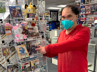 A person wearing a mask and a red shirt, pointing at a display of various collectibles in a store, including Pokémon plush toys and Funko Pop! figures.