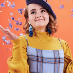 Millicent Simmonds \wearing a beret and tassel and tossing blue petals into the air against an orange background.