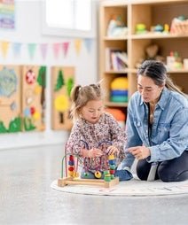 An adult and a child are sitting on the floor, engaging with colorful building blocks in a bright, art-decorated classroom.