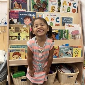 A little girl in a grey and white striped dress posing in front of a bookcase in a classroom.