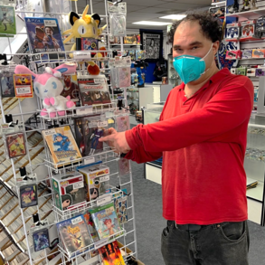 A person wearing a mask and a red shirt, pointing at a display of various collectibles in a store, including Pokémon plush toys and Funko Pop! figures.