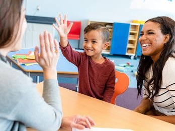 A young child high-fives a teacher across a table in a brightly colored classroom, while another adult smiles beside him, showing an encouraging and cheerful atmosphere.