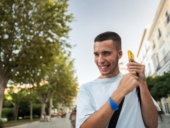 A person smiling and using his smartphone outside with trees and buildings in the background.