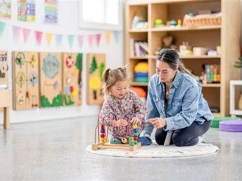 An adult and a child are sitting on the floor, engaging with colorful building blocks in a bright, art-decorated classroom.