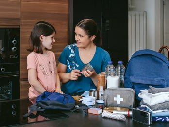 An adult and a child smiling at each other while organizing emergency supplies including water bottles, a first aid kit, and canned food in a kitchen.