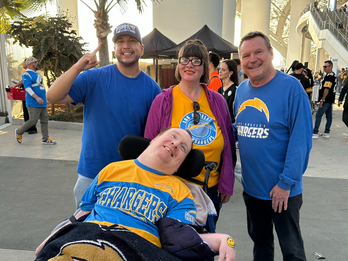 A group of four people wearing San Diego Chargers jerseys posing together outdoors, one person seated in a wheelchair.
