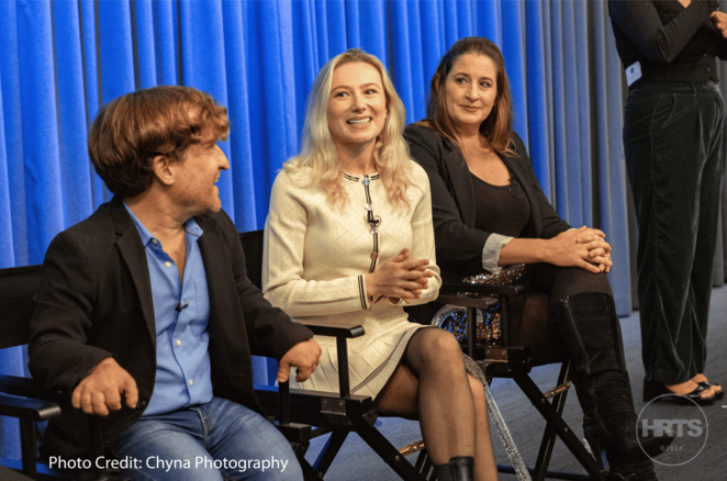 Three individuals are seated side by side at a panel discussion, smiling and engaging with the audience. The setting includes a backdrop with a blue curtain and the logo of HRTS.