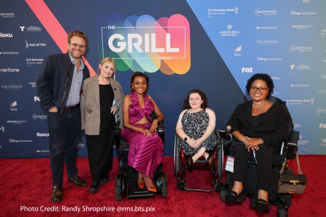 Five individuals posing together on a red carpet at "The Grill" event, with colorful background featuring event logos. Two individuals are seated in wheelchairs.