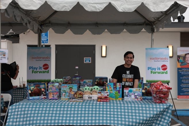 Two individuals exchanging items at a community event, with one person handing over a boxed toy to another who is offering a small item in return. There are tents and other people in the background.