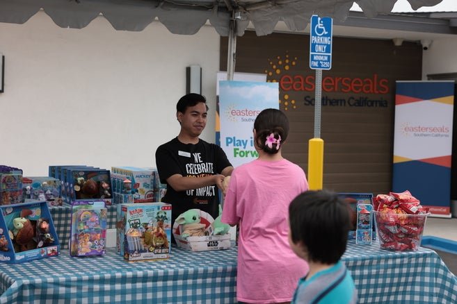 Person standing behind a stall full of toys and games at an Easterseals Southern California event, interacting with a visitor.