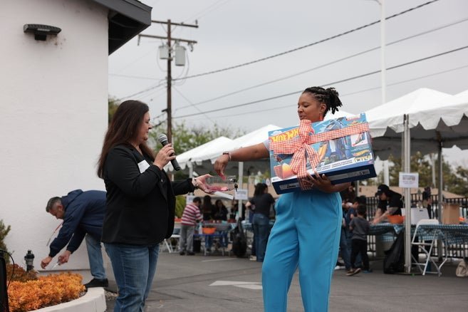 Two individuals exchanging items at a community event, with one person handing over a boxed toy to another who is offering a small item in return. There are tents and other people in the background.