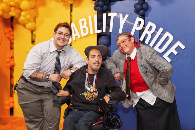 Two Easterseals staff members pose beside a participant in front of a colorful Disability Pride back drop