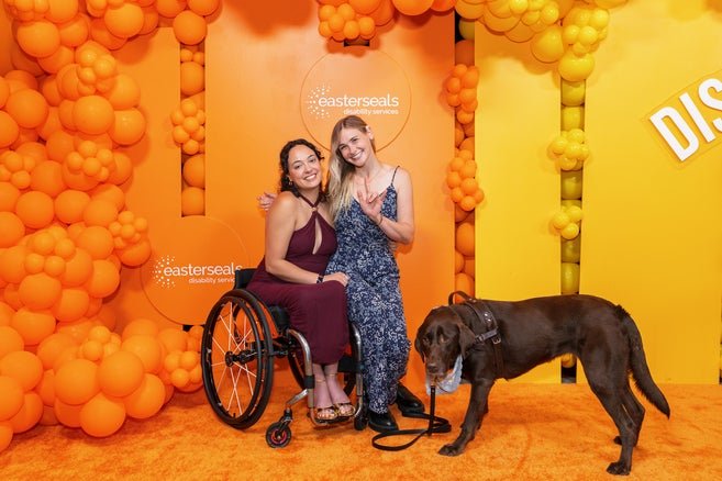 Two women posing in front of a orange and yellow backdrop, one dressed in a burgundy dress sitting in a wheelchair on the left and on the right a woman in a blue dress signing the camera. A service dog on the right is adorably looking at the camera. 