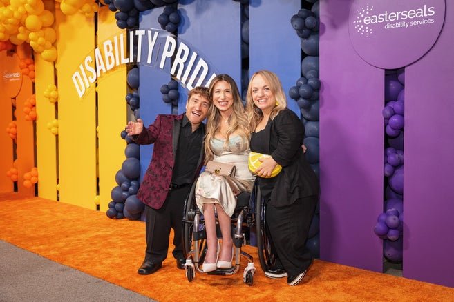 Nic Novicki, Chelsey Hill, and Teale Novicki posing in front of a colorful backdrop that says "disability pride." 
