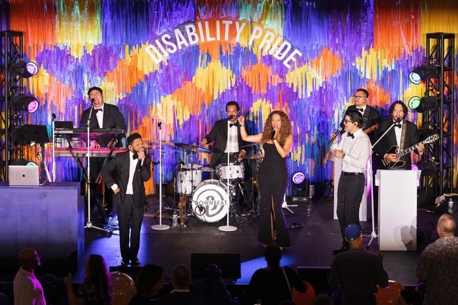 The band on stage performing in front of a purple, blue, orange and yellow fringe backdrop in front of a sign that says disability pride. 