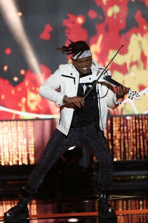 Brian King Joseph performs energetically on an electric violin on stage, with a vibrant fiery backdrop.