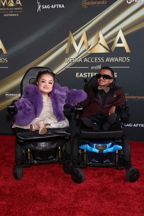 Two attendees at the Media Access Awards, each in an electric wheelchair, posing on the red carpet with a shiny gold backdrop featuring event logos.
