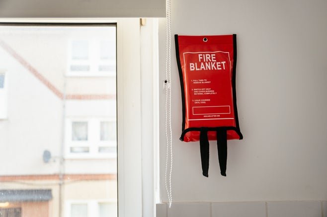 A fire blanket in a red container hangs on a wall near a window, with instructions clearly visible on the front.