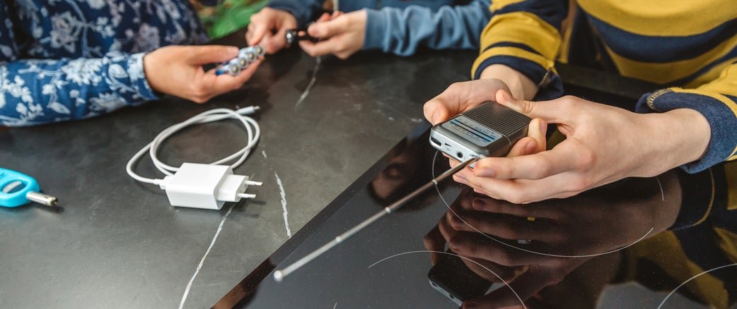 Two individuals sitting at a table working on electronic devices, including a smartphone with tools and a charger cable nearby.