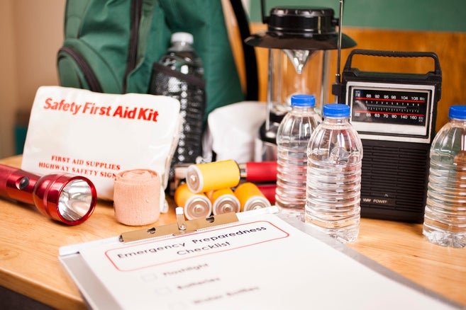 Assorted emergency preparedness supplies on a table, including a first aid kit, bottled water, batteries, a portable radio, a flashlight, and a checklist.