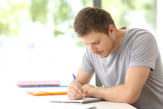 A person sitting at a table writing in a notebook with a pen, in a bright room with windows in the background.