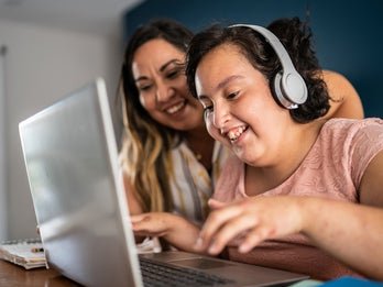 young girl with disability wearing headphones and using a laptop next to adult woman smiling