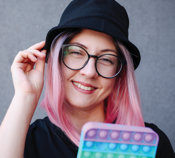 Sarah Hendrickx with pink hair wearing a black bucket hat and black rimmed glasses smiling and holding a sensory toy.